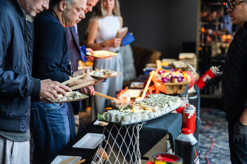 Several people engage in conversation as they stand in line at a buffet, serving themselves sushi from a large platter. The table is decorated with bottles of sauce and various food items, while a woman in the background holds a clipboard.