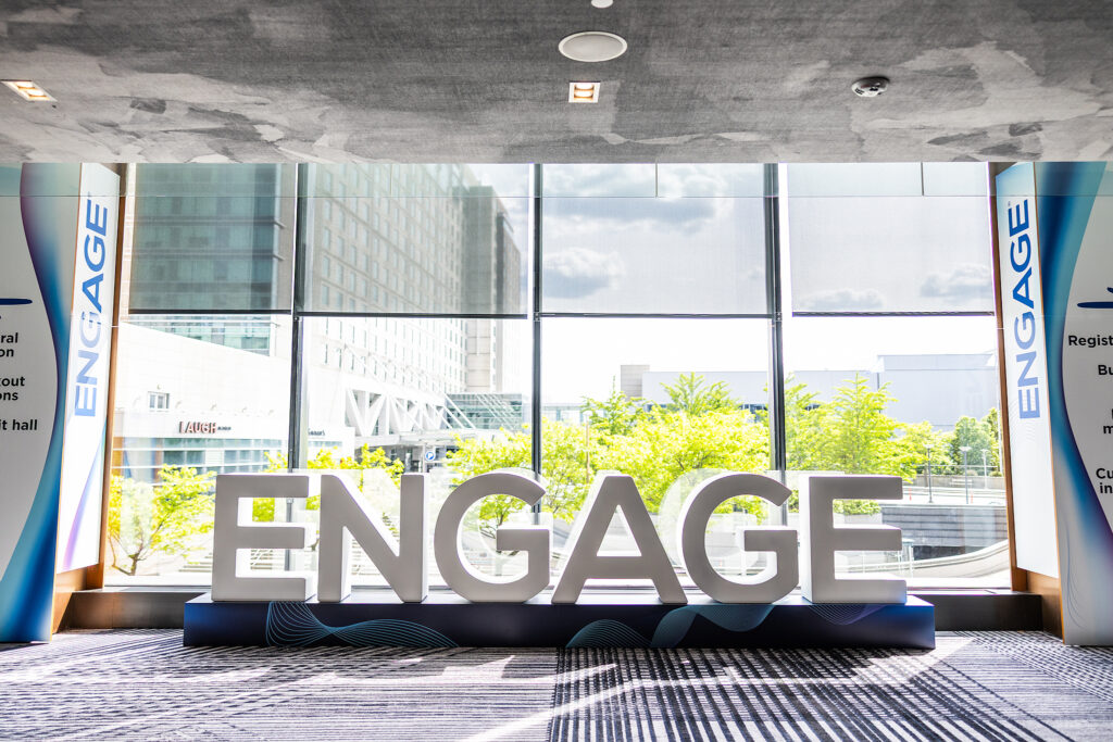 Large, illuminated letters spelling ENGAGE stand boldly in front of tall windows with a sunny cityscape beyond—perfect for capturing the energy of a tradeshow. Blue banners on both sides also read ENGAGE.