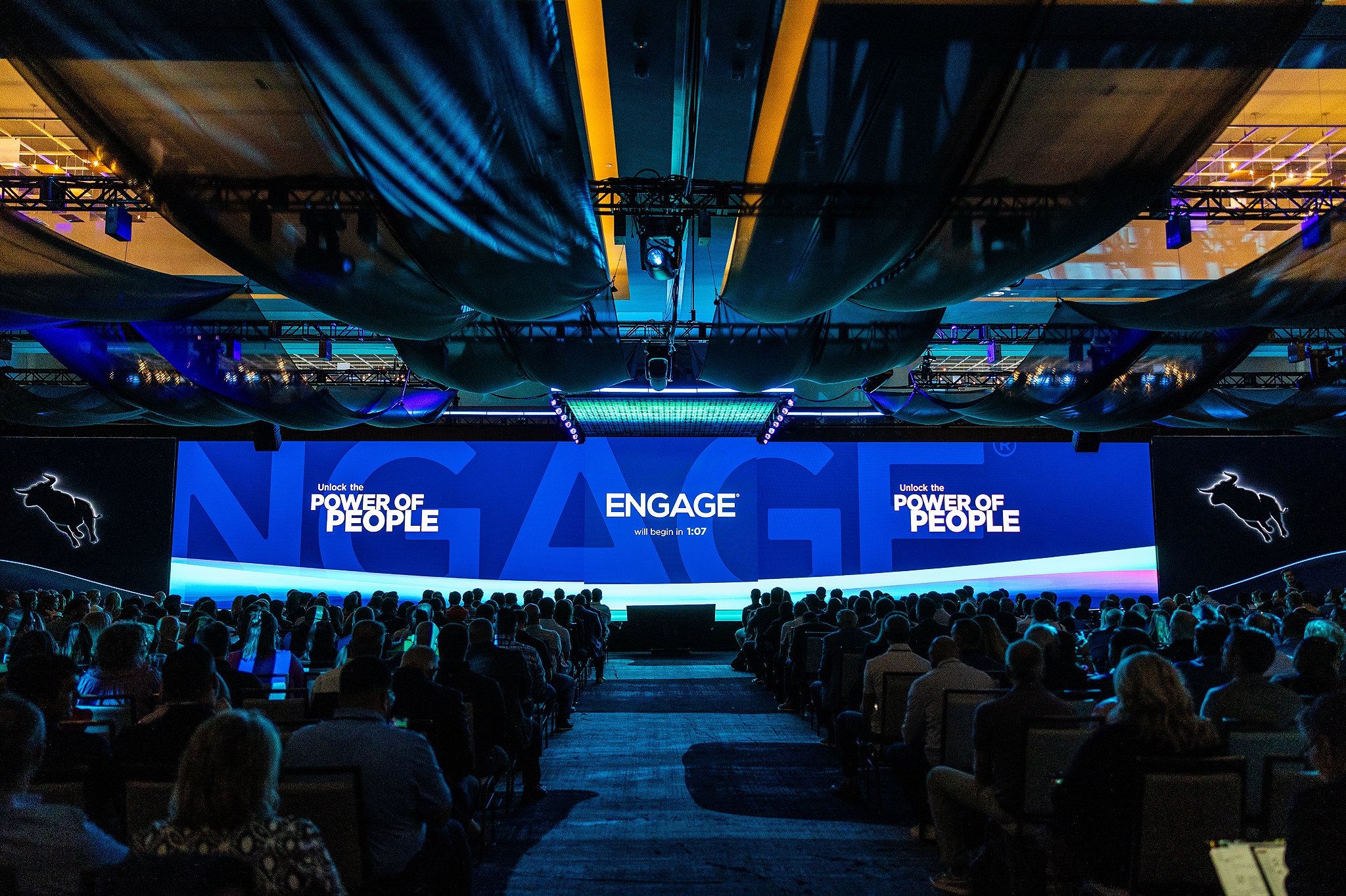 A large audience sits in a dimly lit conference hall facing a wide stage with blue lighting and large screens displaying the words ENGAGE and POWER OF PEOPLE at a tradeshow focused on corporate videography.