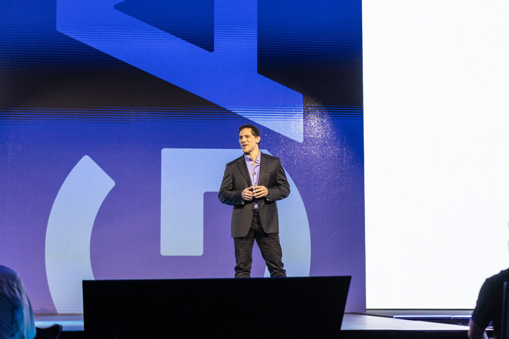 A man in a dark suit and open-collared shirt stands on stage at a Boston tradeshow, speaking to an audience against a blue background with geometric shapes, possibly using a bullhorn to engage the crowd.