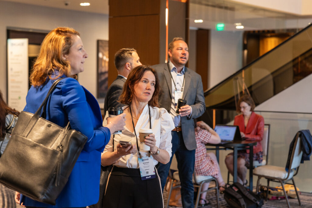 A group of professionally dressed people stand and talk indoors, holding coffee cups at a Boston tradeshow. In the background, others work on laptops at a table near a staircase, suggesting a lively conference or networking event.
