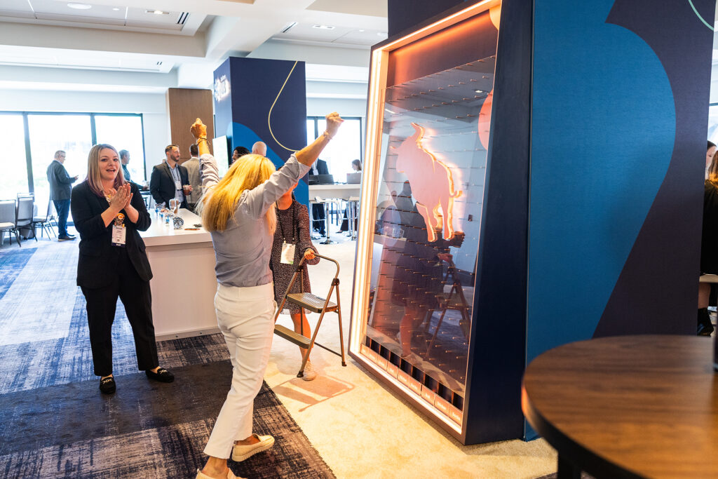 A woman raises her arms in excitement as she plays a large vertical game, engaging the crowd while others watch and clap at a brightly lit corporate event in Boston.