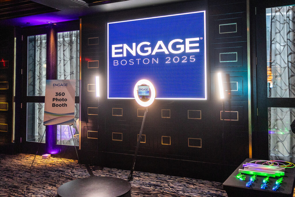 A 360 photo booth setup at an event, featuring a large illuminated ENGAGE Boston 2025 sign, ring light, and a table with colorful glow sticks—perfect for capturing memorable moments with corporate photography in a dimly lit room.