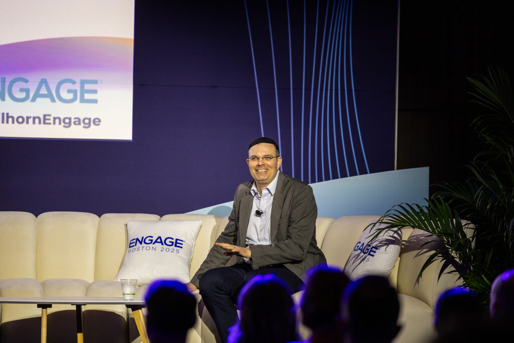 A man in a suit sits on a white couch, smiling and speaking at the ENGAGE Boston 2023 conference. Branded pillows and backdrop highlight the event, while audience members look on—captured with professional corporate videography.