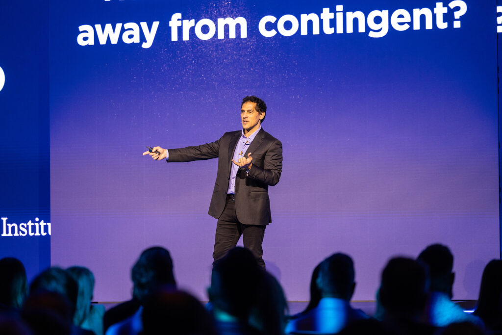 A man in a suit speaks on stage in front of a large blue screen displaying partial text, engaging the Boston audience as he gestures with one hand and holds a device, resembling a digital bullhorn, in the other.