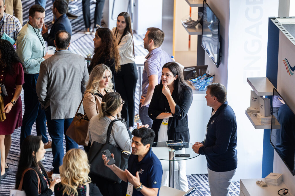 A group of people engage and network at a professional event, standing and talking in small clusters near display booths in a bright, modern indoor space.