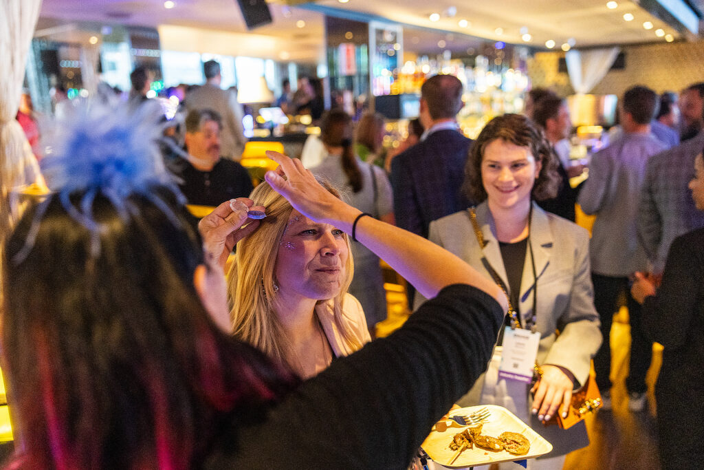 At a lively tradeshow, a woman applies glitter to another woman's forehead while attendees socialize in the festive, busy atmosphere. Nearby, a smiling woman wearing a name badge holds a drink and enjoys the vibrant event.
