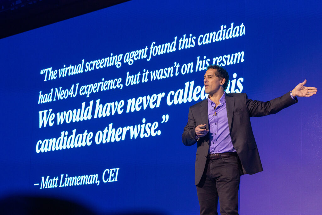 A man in a suit speaks on stage at a Boston tradeshow, in front of a large screen displaying a quote about virtual screening and candidate experience by Matt Linneman, CEI, with a blue background—captured through professional corporate videography.