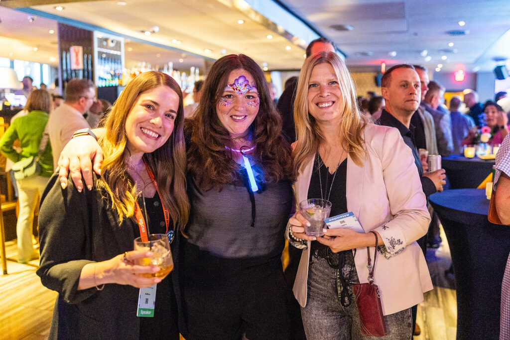 Three smiling women pose together at a lively Boston tradeshow event, holding drinks. One has face paint and a glowing necklace. People engage and mingle in the background, creating a vibrant social atmosphere.