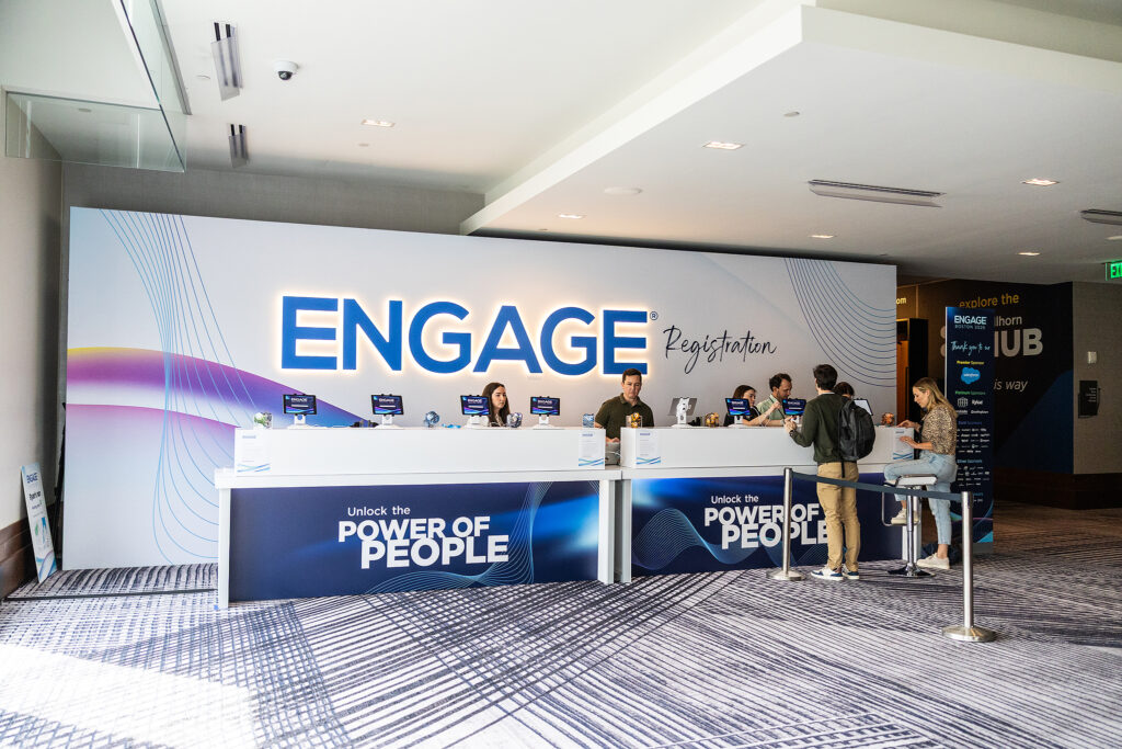 Four people stand at a registration counter under a large ENGAGE Registration sign at this Boston tradeshow, interacting with staff. The modern area features branding and the slogan Unlock the Power of People.