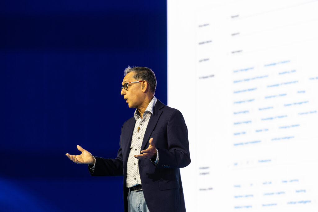 A man in a suit jacket and glasses gestures while speaking on stage at a tradeshow in Boston, with a large, partially visible screen displaying text and tables on a blue and white background—perfect for corporate photography.