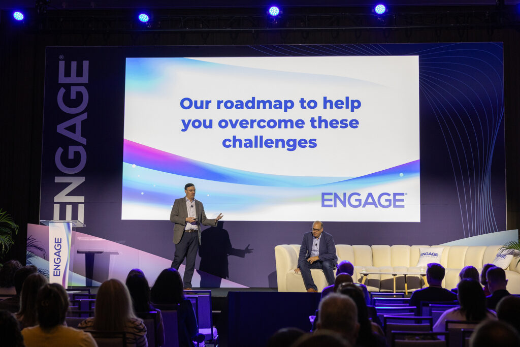 Two men present on stage at the ENGAGE tradeshow, with a large screen behind them displaying, "Our roadmap to help you overcome these challenges." The audience watches from seated rows, capturing a moment perfect for corporate videography.