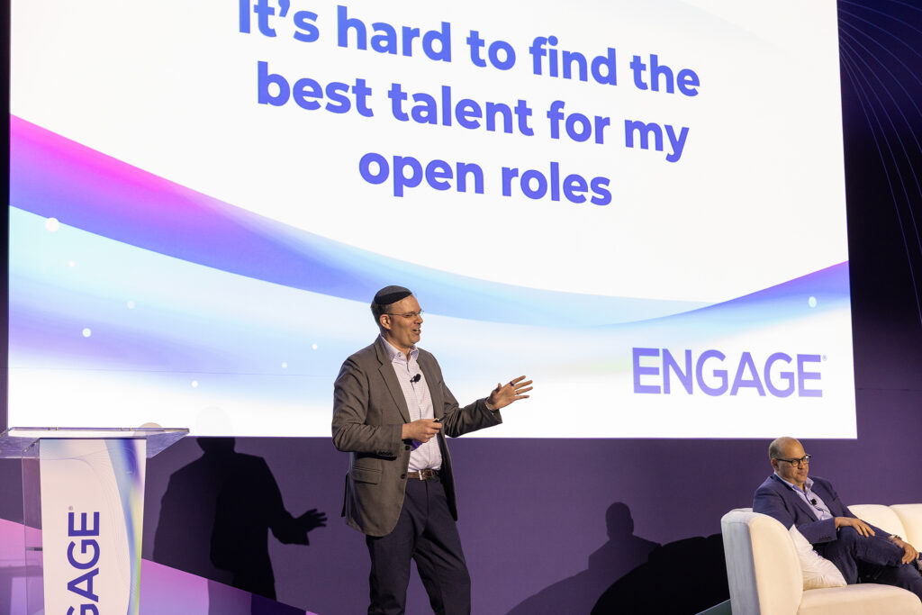 A man stands on stage speaking near a seated man, with a large screen behind them displaying “It’s hard to find the best talent for my open roles” and “ENGAGE” in the corner, capturing a moment of impactful corporate videography.