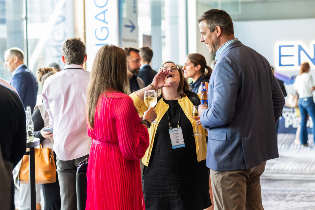 A group of people in business attire mingle at a Boston conference. In the center, a woman laughs with her hand raised while holding a drink—captured perfectly through corporate photography. Others chat nearby, with large signs in the background.