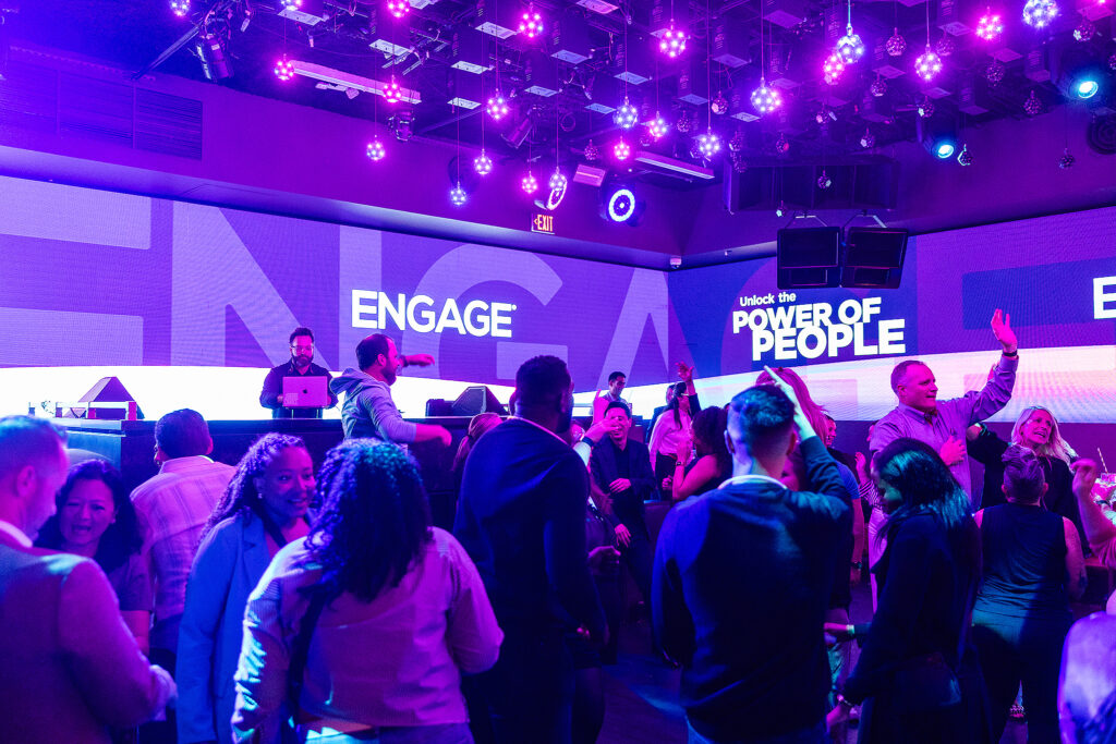 A crowd of people dances and socializes in a vibrant club with purple lighting, like an energetic tradeshow afterparty. A DJ plays music on stage in front of large screens displaying the words ENGAGE and Unlock the POWER OF PEOPLE.