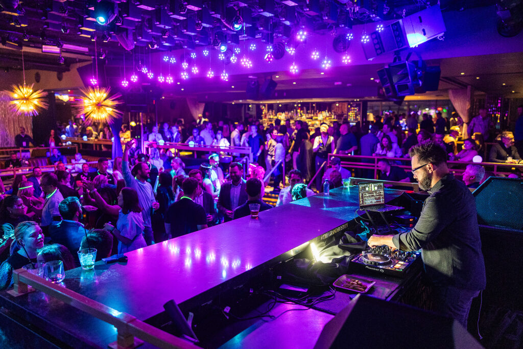 A crowded nightclub with colorful purple and blue lighting, people dancing and socializing, and a DJ playing music at a booth in the foreground. The lively atmosphere makes it easy to engage with others—perfect for dynamic corporate photography.