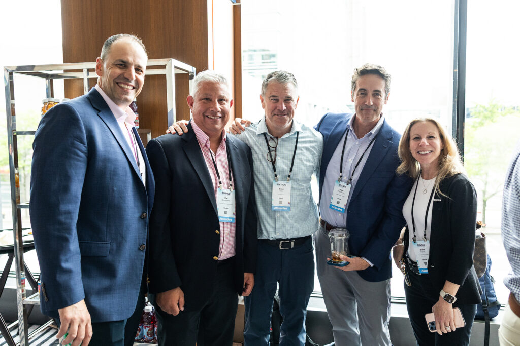 Five people in business attire stand closely together, smiling at the camera at a tradeshow networking event. They wear name badges and are indoors with large windows in the background, capturing the spirit of corporate photography.