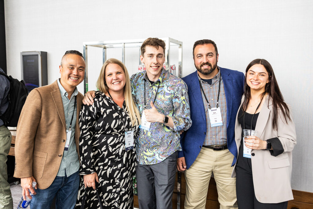 Five people stand together indoors in Boston, smiling at the camera. Dressed in business casual attire with conference badges, one gives a thumbs up while another holds a drink—a perfect moment for corporate videography.