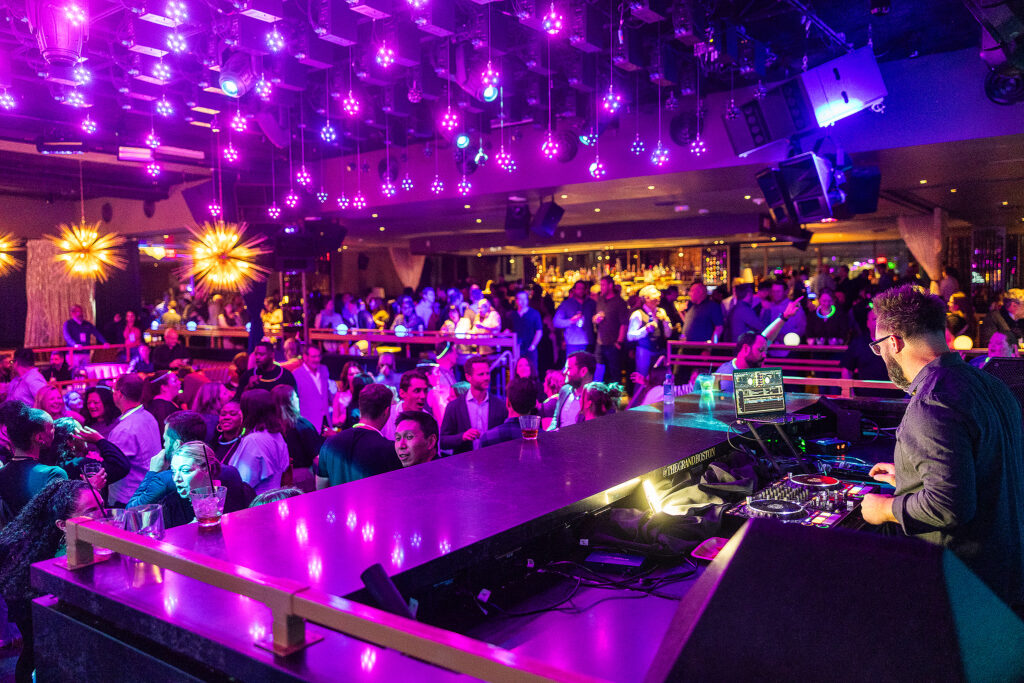A crowded nightclub with colorful purple lights, a DJ playing music at a booth, and people socializing, dancing, and sitting at tables in a lively atmosphere reminiscent of a tradeshow afterparty.
