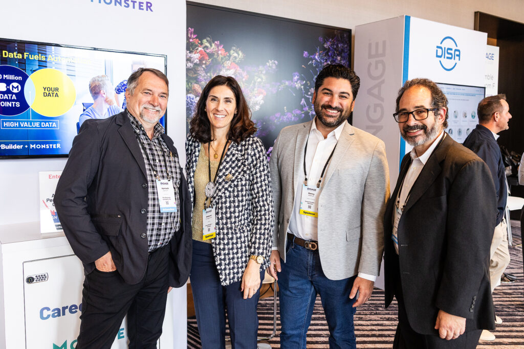 Four people, three men and one woman, stand together smiling at a professional event in Boston. They wear business attire and conference badges, with exhibition booths and digital screens featured in this corporate photography scene.