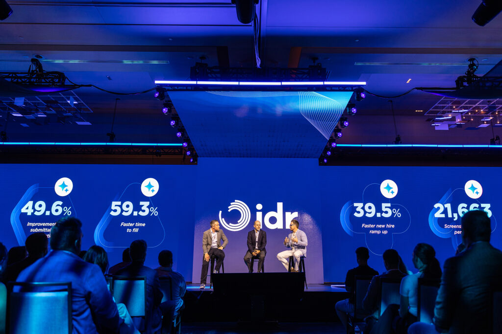 Three speakers sit on stage in Boston at a conference, engaging the audience as statistics and percentages are highlighted in blue graphics behind them under the idr logo.