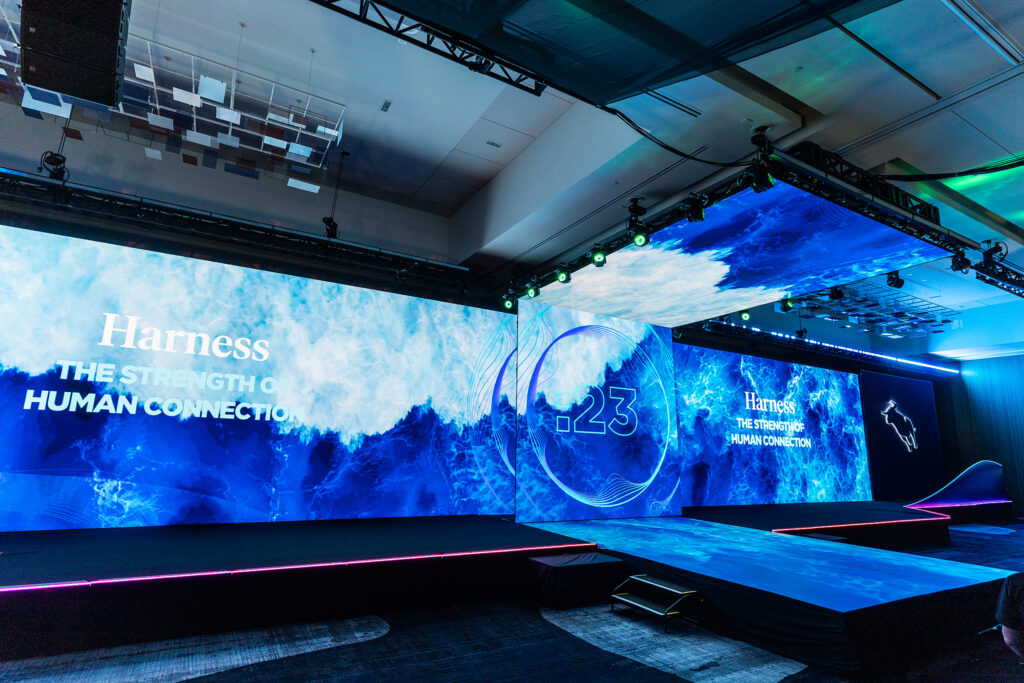 A large, modern stage setup with blue lighting and ocean-themed visuals displays the words Harness the strength of human connection and 23 on multiple wide screens at a Boston tradeshow conference hall.