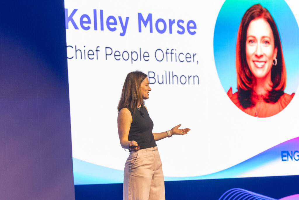 A woman stands on stage presenting at a tradeshow in Boston, gesturing with her hands in front of a large screen displaying Kelley Morse, Chief People Officer at Bullhorn, alongside her portrait photo.
