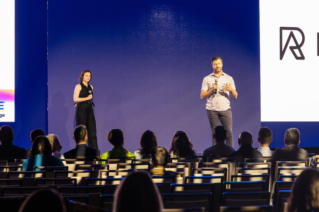 Two people stand on stage presenting to an engaged audience in a dimly lit Boston venue. The woman on the left holds a microphone, while the man speaks. A large screen with a logo glows behind them, capturing the essence of corporate videography.