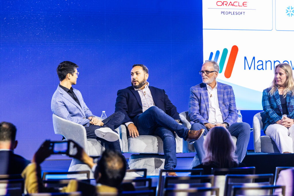 Four people sit on stage in a panel discussion at a Boston tradeshow, engaging with the audience. Three men and one woman are seated in armchairs with a blue background and company logos displayed behind them. An audience member takes a photo.