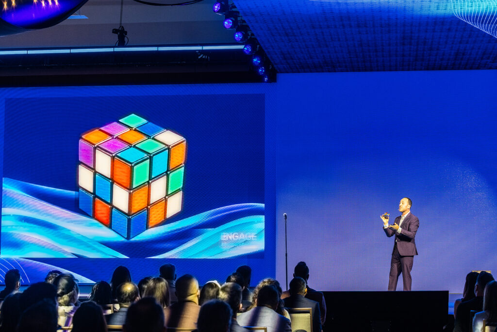 A speaker in a suit gestures on stage at a tradeshow, engaging the audience as a large screen displays a colorful digital Rubik’s Cube and blue wave patterns in the background.