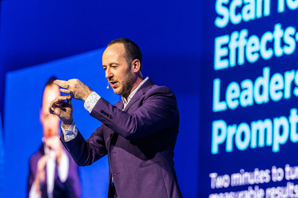 A man in a purple suit gestures while speaking on stage in front of a large blue screen displaying white text about leadership and prompt effects, showcasing the power of corporate videography to engage audiences. A blurred image of him appears on another screen behind.