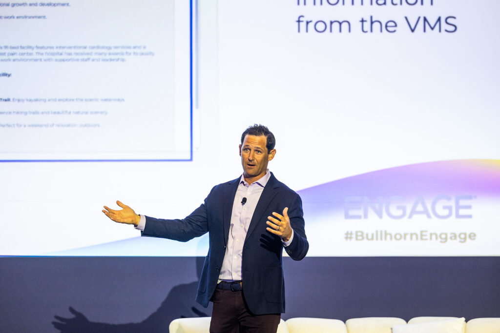 A man in a suit gestures while speaking on stage at a Boston conference, with the screen behind him displaying text and the words ENGAGE #BullhornEngage. White chairs are visible in the foreground, capturing a moment of dynamic corporate photography.
