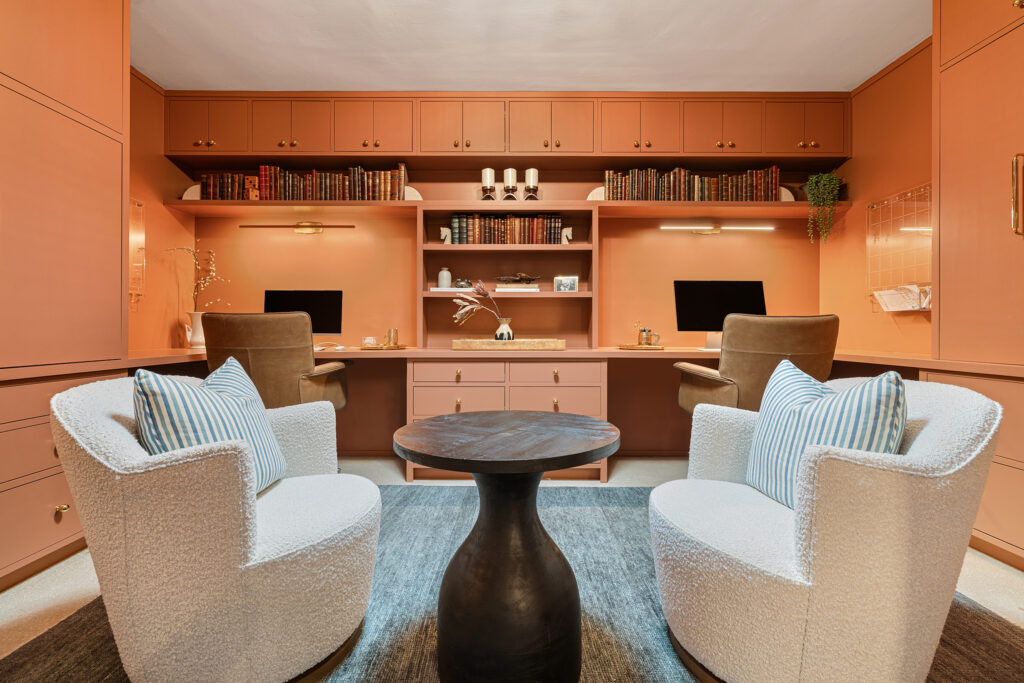A stylish home office design with peach built-in cabinets, two desk areas with computers, brown office chairs, and designer white swivel chairs around a round black table on a gray rug. Shelves hold books and decor.