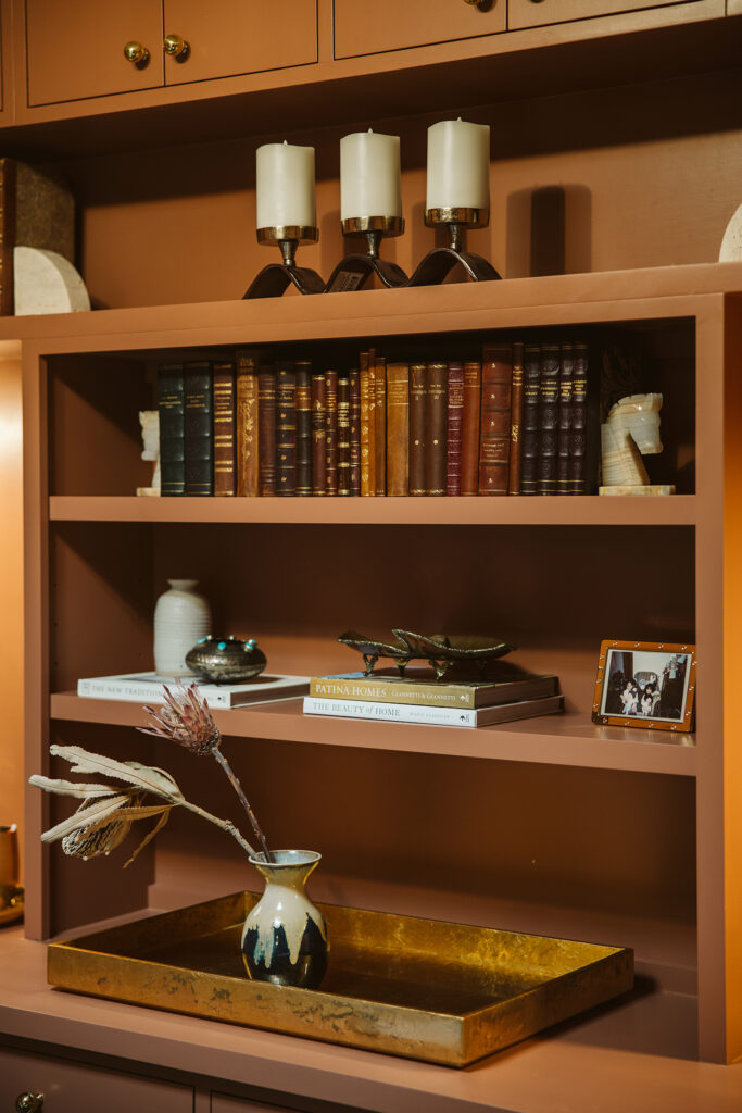 A designer bookshelf with candles, vintage books, horse-head bookends, vases, decorative objects, a framed photo, and stacked design books—including Patina Homes and The Beauty of Home—elevates your office on a gold tray.
