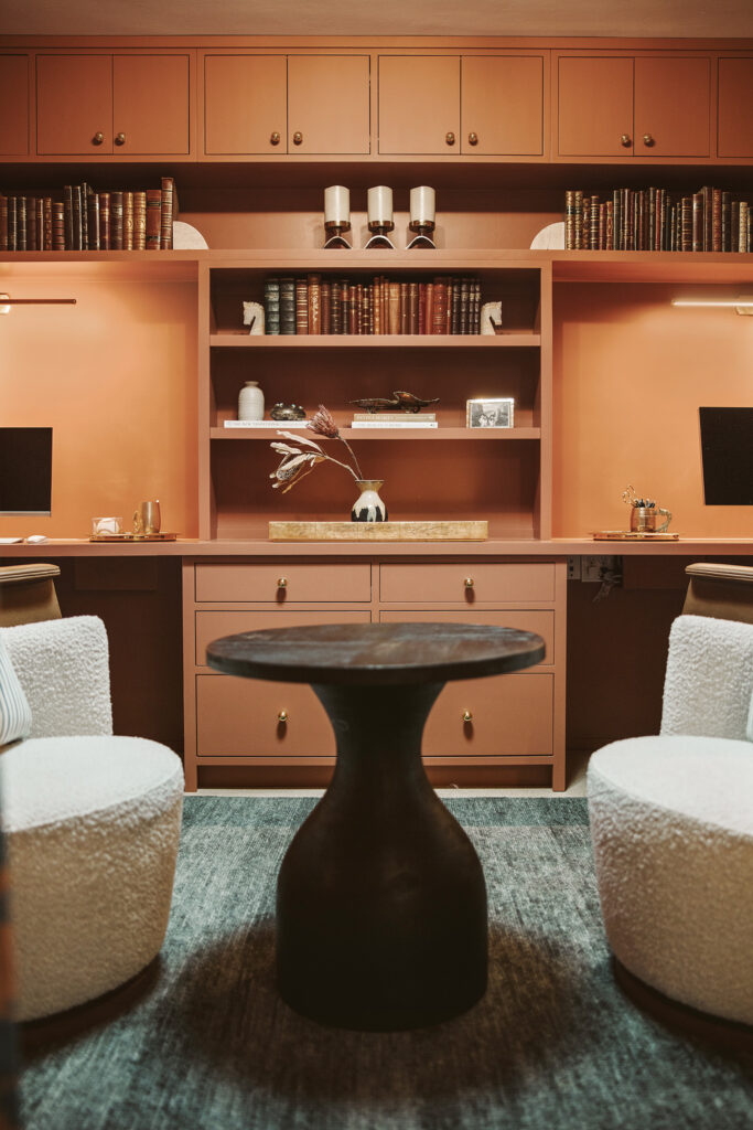 A cozy Kim Wolfe office design featuring two white chairs, a round dark wood table, orange built-in shelves filled with books and decor, dual computer monitors, and warm ambient lighting.