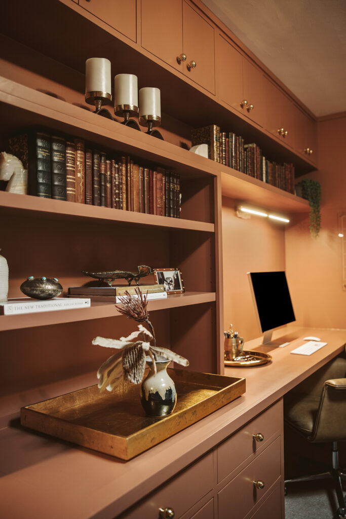 A stylish home office designed by Kim Wolfe, featuring terracotta-colored shelves and cabinets, books, decorative items, a tray with a vase, and a neatly arranged desk with a desktop computer and brown leather chair.