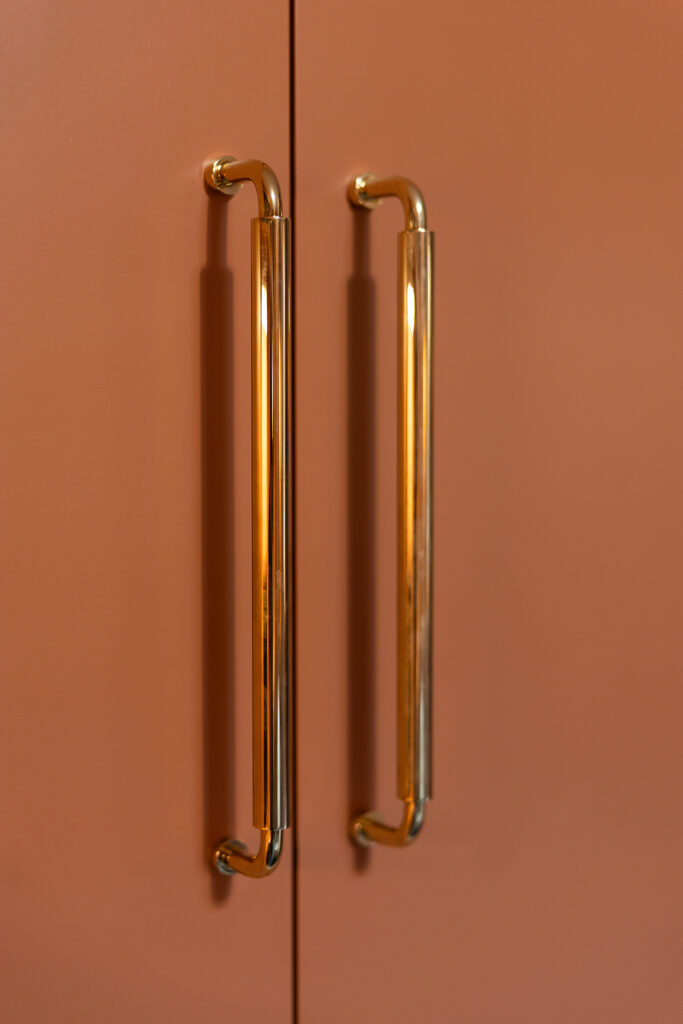 Close-up of two vertical gold cabinet handles mounted on matte, terracotta-colored doors—a stylish design detail inspired by Kim Wolfe, perfect for a chic office space.