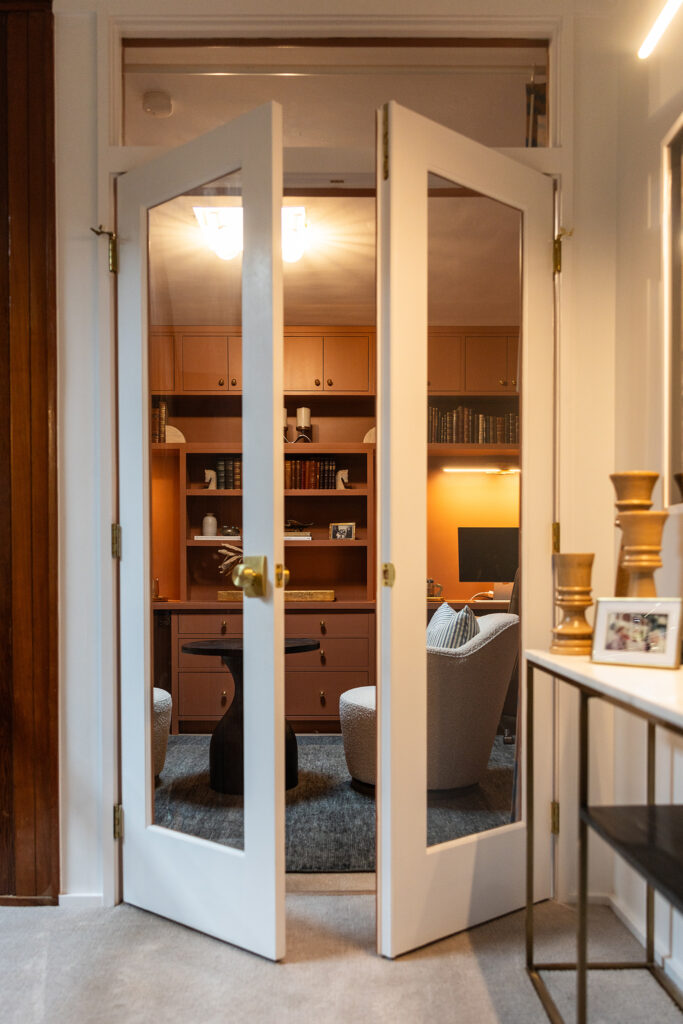 Glass double doors open to reveal a cozy home office designed by Kim Wolfe, with shelves of books, a desk with a computer monitor, two modern chairs, and warm lighting. A console table with curated decor sits in the foreground.