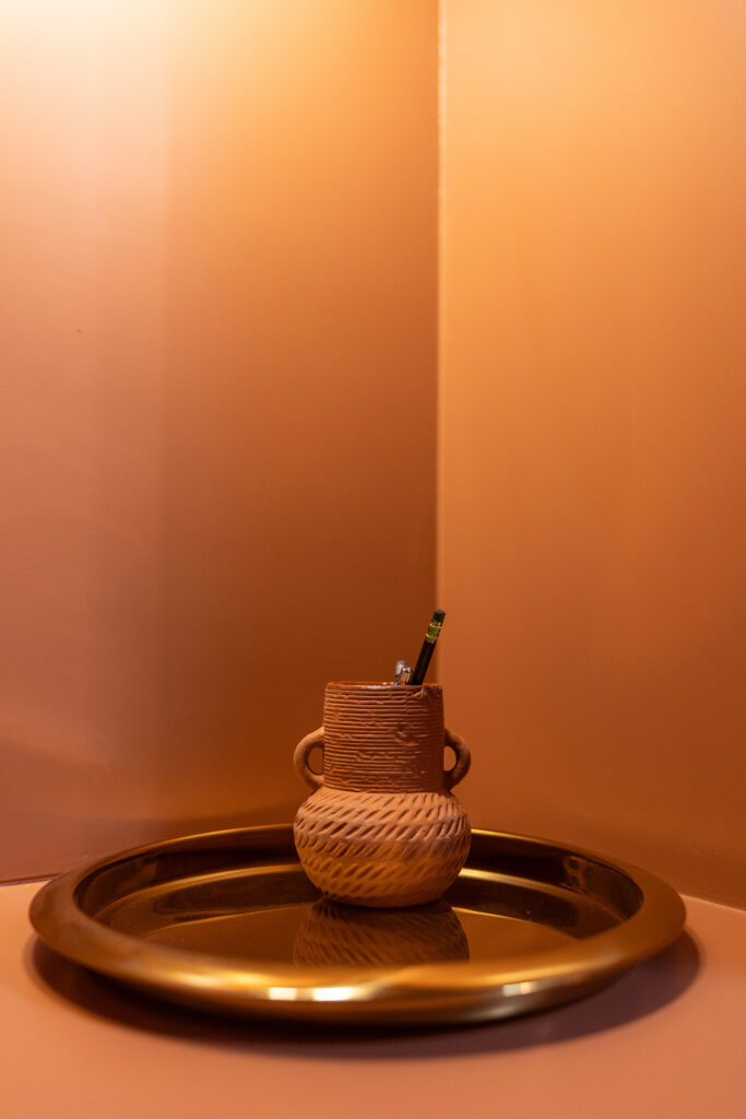 A pencil holder shaped like a decorative clay pot with handles, designed by Kim Wolfe, sits on a round, reflective brass tray in a corner with warm, orange lighting. A pencil and a pen are inside the holder.