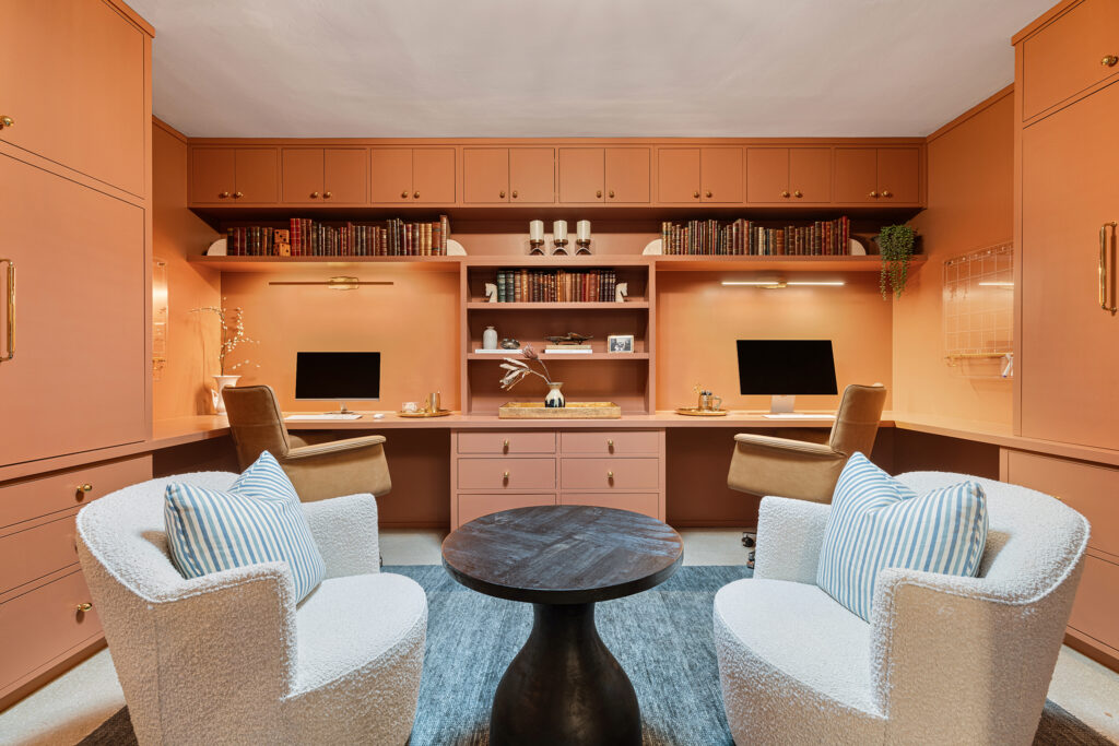 A stylish home office showcases designer flair with two desks and computers, peach cabinetry, bookshelves, and gold accents. Two white lounge chairs with striped pillows face a round dark coffee table on a gray rug.