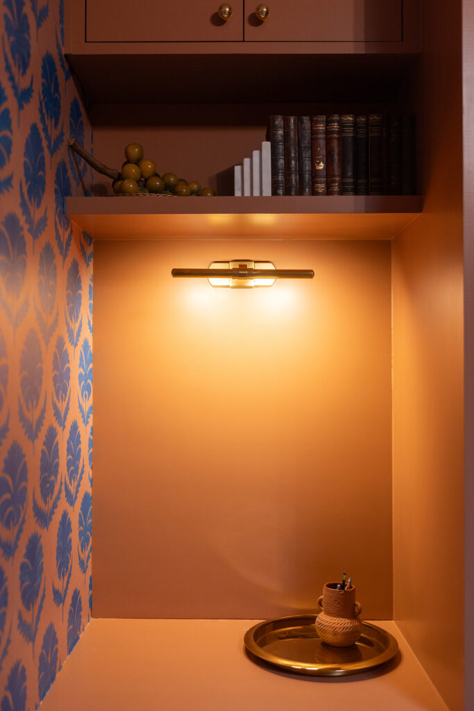 A small office alcove with an orange wall, designed in Kim Wolfe’s style and lit by a wall lamp. A gold tray with pens in a holder sits on the surface, while books and artificial grapes rest on a shelf above. Blue patterned wallpaper decorates the side wall.