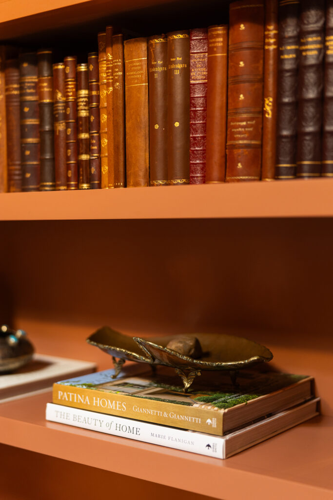 A bookshelf with antique leather-bound books on the upper shelf and two modern design books stacked below, topped with a decorative item shaped like a large bronze leaf—perfect for any designer’s curated space.