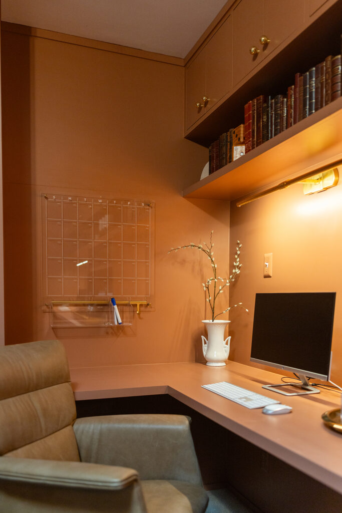 A modern home office designed with tan walls and cabinets, a leather chair, sleek desk setup, white vase with branches, books on a shelf, and a wall-mounted organizer—an inspiring space worthy of designer Kim Wolfe.