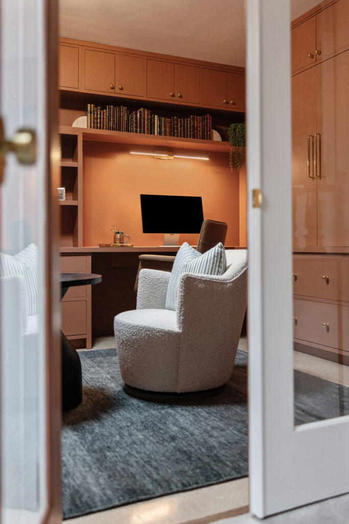Modern home office with warm peach walls and built-in shelving, designed in the style of Kim Wolfe, featuring a desk with a computer monitor, a cozy swivel chair with striped pillows, and a gray rug—all viewed through glass-paneled doors.