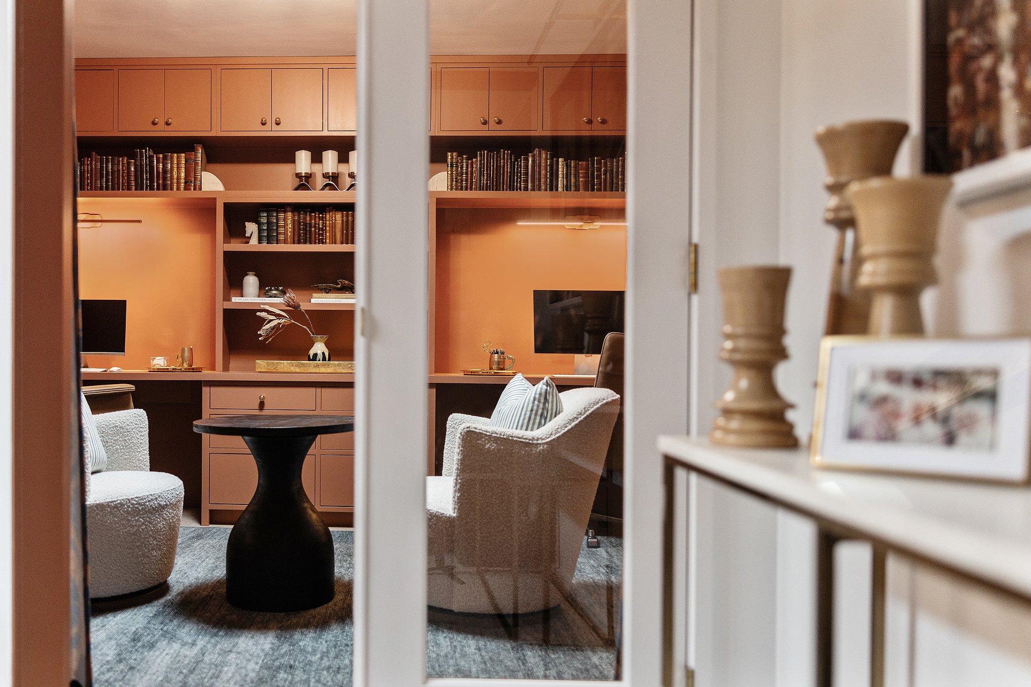 A cozy home office by designer Kim Wolfe features two white armchairs around a round table, built-in shelves filled with books, two computer monitors, and warm peach-colored walls, all viewed through elegant glass double doors.