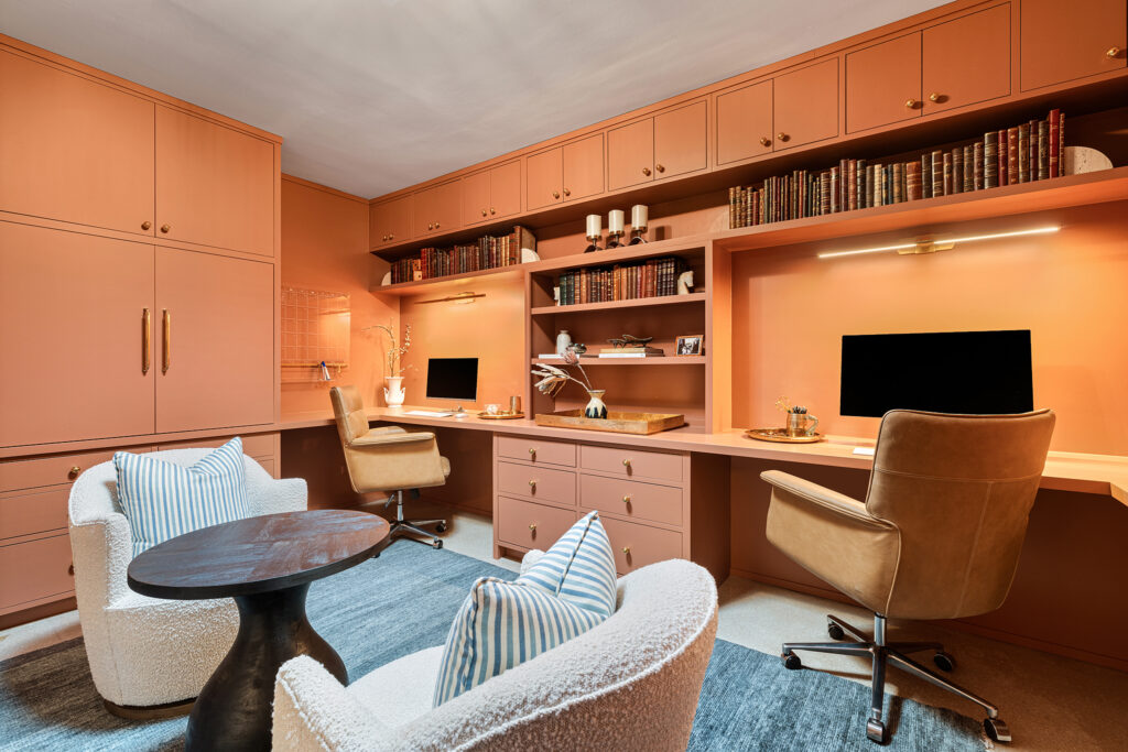 A stylish home office by designer Kim Wolfe features peach cabinetry, built-in desks, dual monitors, leather chairs, bookshelves filled with books, and a seating area with two armchairs, a small round table, and a blue rug.