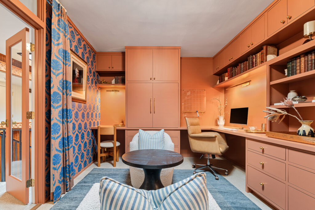 A cozy home office with peach-colored cabinets, built-in desk, bookshelves, and inviting Kim Wolfe-inspired design accents. Blue patterned curtains frame striped pillows on a white and blue rug, with warm lighting creating a welcoming vibe.
