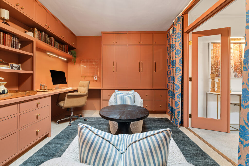 A stylish home office inspired by Kim Wolfe’s design, featuring terracotta built-in cabinets, a desk and shelves, computer, beige office chair, round black coffee table, striped armchairs, and blue patterned curtains. The room is well-lit and cozy.