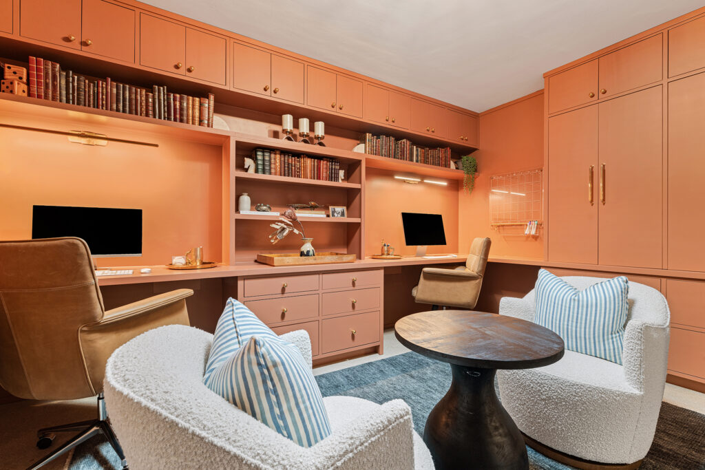 A stylish home office designed by Kim Wolfe features peach-colored built-in shelves, two desks with computers, tan chairs, and a round black table between two white armchairs with blue-striped pillows. Shelves display books and decor items.