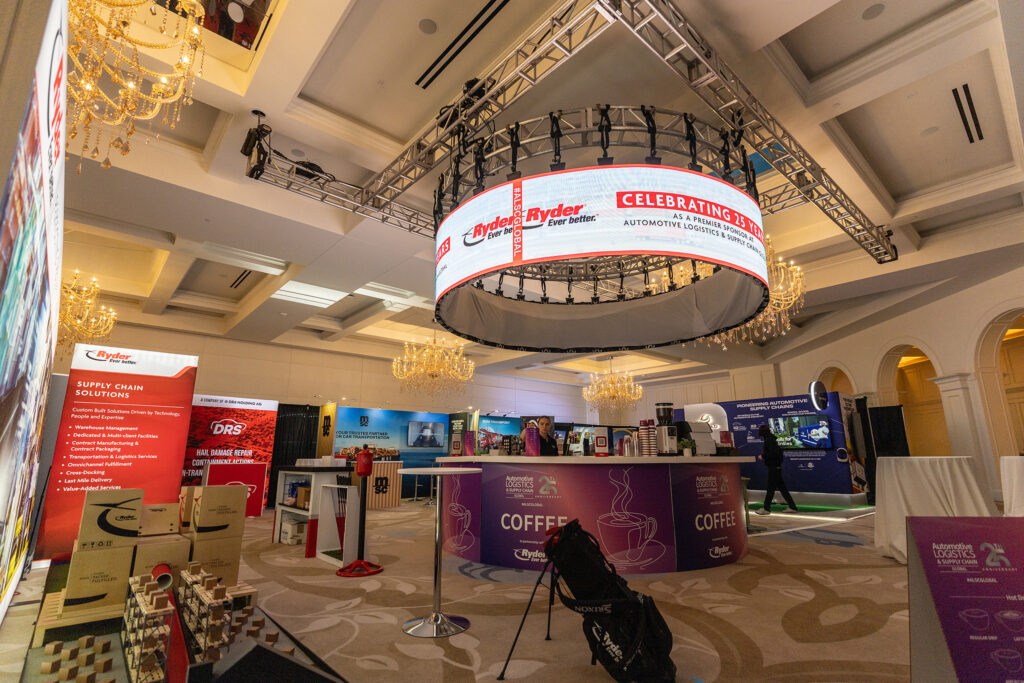 A brightly lit corporate conference room with exhibition booths, a large circular LED display overhead, and ALCS banners; chandeliers hang from the ceiling at the Automotive Logistics and Supply Chain Global event.